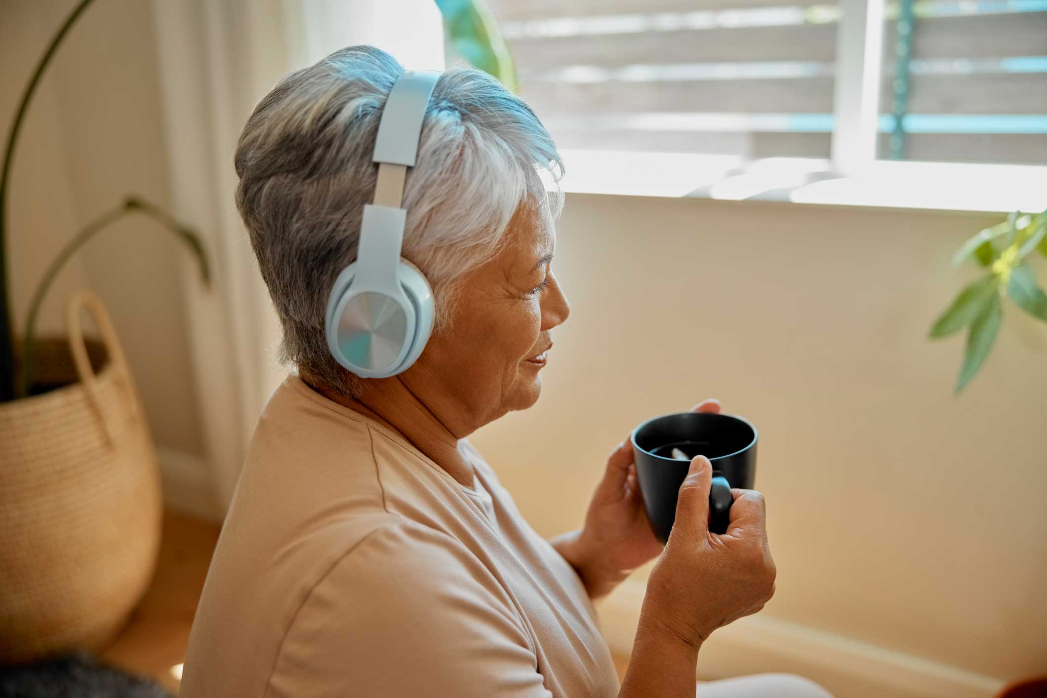 senior woman in her living room drinking coffee and listening to music with headphones