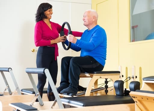 Cheerful Hispanic woman fitness trainer controlling positive elderly man performing exercises with Magic Circle in pilates studio